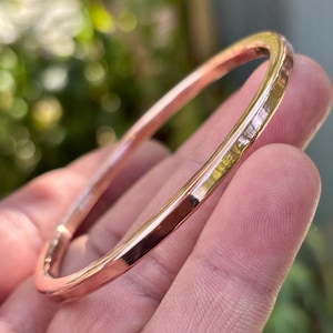 May include: A hammered copper bangle bracelet. The bracelet has a rounded square shape and a textured surface. The copper has a warm, reddish-brown color with highlights of gold. The bracelet is shown against a blurred green background.