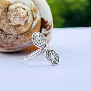 Könnte beinhalten: Ein silberfarbener Ring mit einem doppelten Spiraldesign. Der Ring hat zwei gewundene Spiralen, die durch ein schmales Band verbunden sind. Der Ring liegt auf einer weißen Oberfläche mit einer Muschel im Hintergrund.