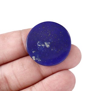 May include: A circular, polished lapis lazuli gemstone cabochon featuring a deep royal blue colour with scattered golden pyrite flecks and small white calcite inclusions across its smooth, flat surface, presented in a round, disc-like shape.