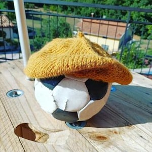 May include: A worn soccer ball with black and white panels is topped with a mustard yellow knitted beret. The beret is made of a textured yarn. The ball sits on a wooden spool in the sunlight.