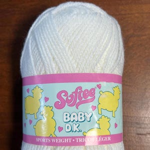Puede incluir: Una madeja de hilo blanco roto con una etiqueta rosa y turquesa. La etiqueta dice "Softee BABY D.K." con ilustraciones de ovejas amarillas y las palabras "Sports Weight • Tricot Léger".