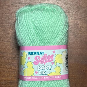 Puede incluir: Una madeja de hilo verde menta Bernat Softee Baby D.K. La etiqueta es rosa y blanca con el nombre de la marca, el tipo de hilo e ilustraciones de ovejas amarillas. El hilo es de peso deportivo y está etiquetado como "Tricot Léger".