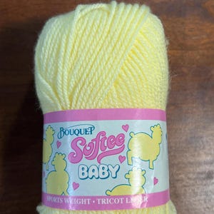 May include: A skein of soft yellow yarn labeled "Bouquet Softee Baby." The label features a pink border with yellow sheep illustrations and the words "Sports Weight - Tricot Laine."