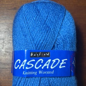 Op de afbeelding: Een bol blauw breiwol met een blauw etiket. Het etiket leest "Hayfield CASCADE Knitting Worsted". Het garen heeft een levendige blauwe kleur, perfect voor breiprojecten.