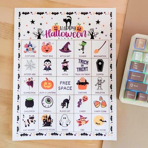 May include: A Halloween bingo card featuring illustrations of pumpkins, witches, and ghosts. The card has the text "Happy Halloween Bingo" at the top. The background is light, and a keyboard is partially visible. The card is designed for a Halloween party.