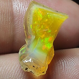 May include: A vibrant, translucent opal gemstone with a mix of yellow, orange, and green internal colors. The rough-cut stone is held between fingers against a dark background, showcasing its natural, irregular shape.