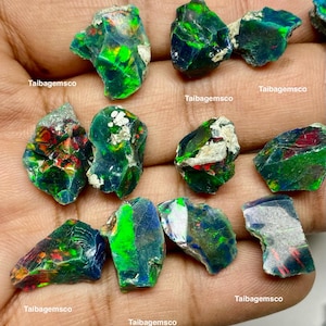 May include: A collection of raw, dark-toned opals featuring vibrant flashes of green, red, and blue play-of-color. Each irregular, unpolished gemstone displays a unique surface texture with visible inclusions and the text Taibagemsco overlaid.