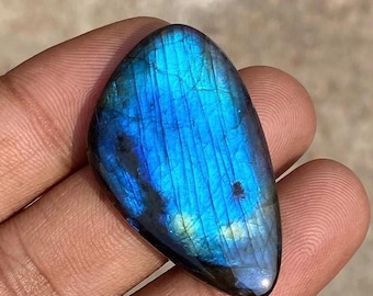 May include: A polished, teardrop-shaped labradorite gemstone. The stone displays a vibrant play of color, with iridescent blues and greens. The surface has a smooth, glossy finish, and the stone is held in a hand.