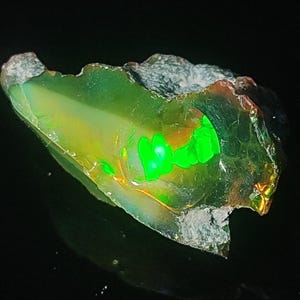 May include: A vibrant, raw opal gemstone with a striking display of color. The stone features a translucent, green body with flashes of bright green and yellow. The rough edges and natural form of the opal are visible.