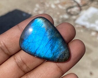May include: A polished, triangular labradorite gemstone. The stone displays a vibrant blue iridescence with streaks of darker blue and gray. The gem is held in a hand, showcasing its natural shape and color variations.