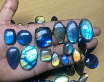 May include: A collection of polished labradorite gemstones in various shapes and sizes, including oval, teardrop, and rectangular. The stones display iridescent blue, green, and gold flashes. Some are held in a hand, while others are scattered.