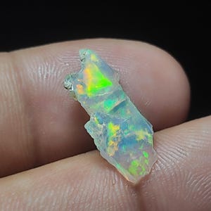 Puede incluir: Fragmento de ópalo iridiscente en bruto. El ópalo exhibe destellos de color verde, amarillo y azul. La piedra se sostiene entre los dedos contra un fondo oscuro, resaltando su forma natural y sin pulir.