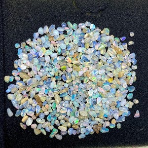 May include: A collection of small, polished opal gemstones in various shapes and sizes. The opals display a range of colors, including blue, green, white, and hints of yellow and orange. The gemstones are scattered on a black surface.