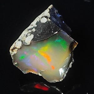 May include: A rough-cut opal gemstone with a dark, textured exterior and a vibrant, iridescent interior displaying flashes of green, orange, and yellow. The opal is set against a black background, highlighting its natural beauty.