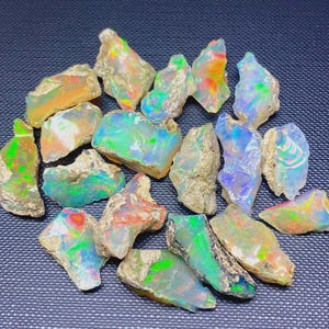 Peut inclure: Un ensemble de pierres d'opale brutes, présentant une variété de couleurs, notamment le vert, le bleu, l'orange et le jaune. Les opales ont une texture brute et naturelle, sur fond sombre.