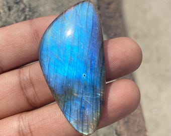 May include: A polished, teardrop-shaped labradorite gemstone. The stone displays a vibrant play of color, with iridescent blues and greens. The surface has natural lines and patterns, adding to its unique appearance. The gemstone is held in a hand.