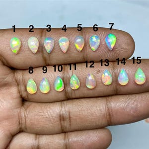 May include: Fifteen pear-shaped opals in various colours, including white, yellow, green, and pink, are displayed. Each opal is numbered from 1 to 15. The opals are held in a hand, showcasing their unique iridescence and natural beauty.