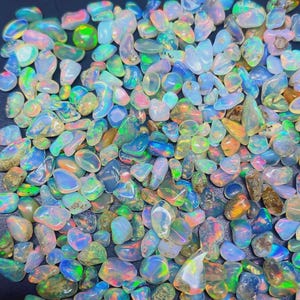 Può includere: Una collezione di pietre preziose di opale lucide di varie forme e dimensioni. Gli opali mostrano una gamma di colori, tra cui blu, verde, arancione e rosa, con lampi iridescenti. Le pietre preziose sono sparse, creando un'esposizione strutturata e colorata.