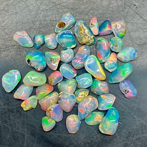 May include: A collection of polished opal gemstones. The opals display a variety of colors, including blue, green, orange, and pink, with iridescent flashes. The stones have a smooth, rounded shape and are scattered across a dark surface.