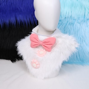 May include: A white faux fur collar with a pink bow and paw print accents. The collar is displayed on a mannequin head, with blue and black faux fur in the background. The collar is designed to be worn around the neck.