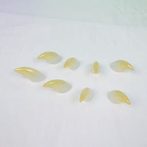 May include: A collection of eight translucent, claw-shaped objects with a gradient of yellow to white. The objects are arranged on a white surface, with varying sizes and orientations, suggesting a set of decorative or artistic elements.