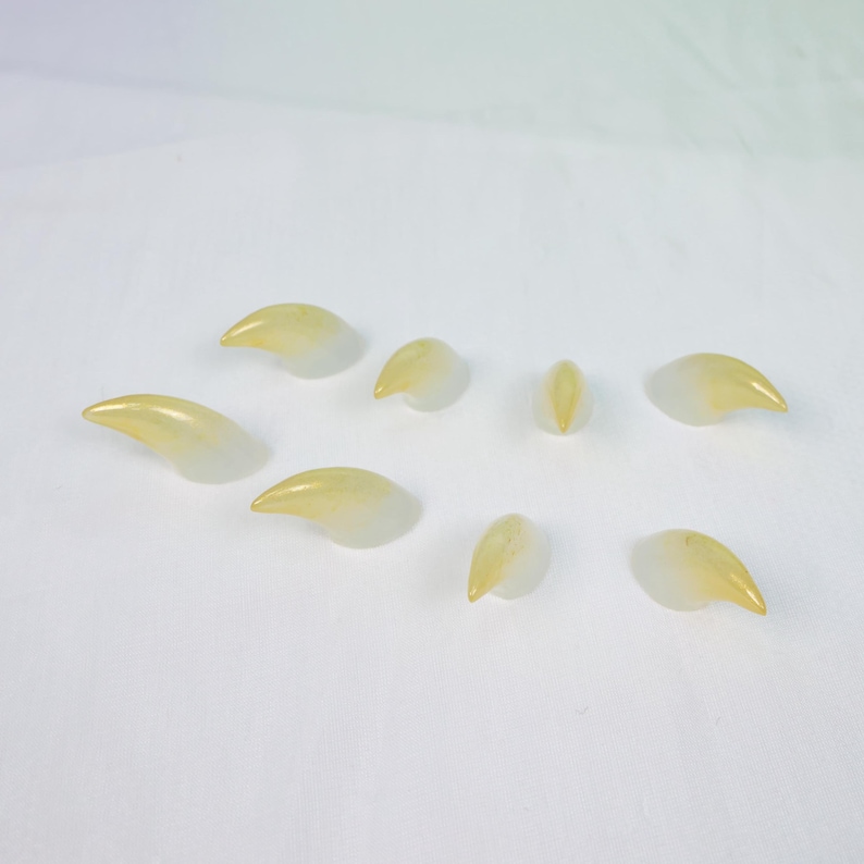 May include: A collection of eight translucent, claw-shaped objects with a gradient of yellow to white. The objects are arranged on a white surface, with varying sizes and orientations, suggesting a set of decorative or artistic elements.