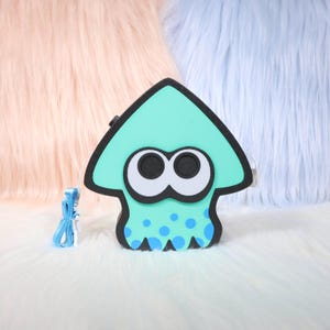 May include: A teal squid-shaped accessory with black trim and large, round eyes. The design includes blue polka dots and a matching blue strap. The background features fluffy pink and blue textures.
