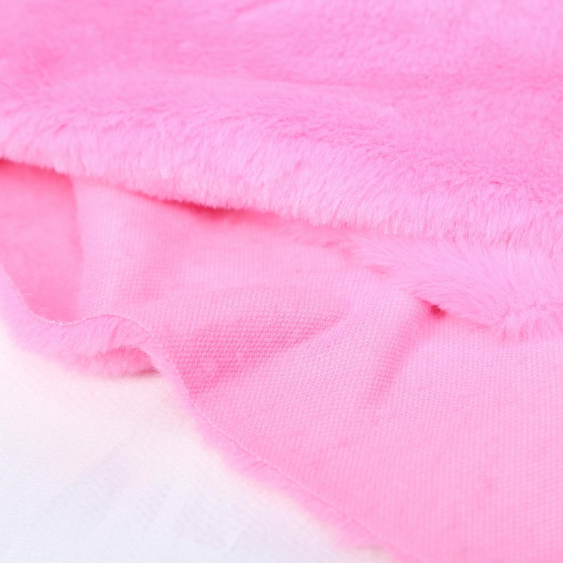 May include: Close-up of pink faux fur fabric with a soft, plush texture. The fabric is layered over a smooth, lighter pink material, creating a contrast in textures. The image highlights the vibrant color and tactile qualities of the materials.