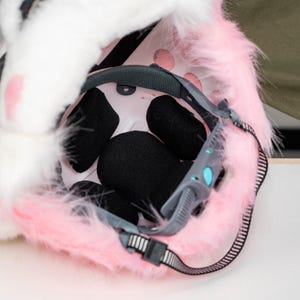 May include: A protective helmet interior featuring a light pink faux fur exterior trim. The inside of the helmet is lined with black foam padding, a grey adjustable suspension system, and a grey chin strap with a plastic buckle closure mechanism.