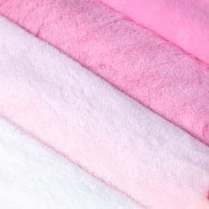 May include: Close-up of soft, plush fabric in shades of pink and white. The textures vary from light to dark pink, creating a gradient effect. The material appears fluffy and inviting, ideal for crafting or home decor projects.