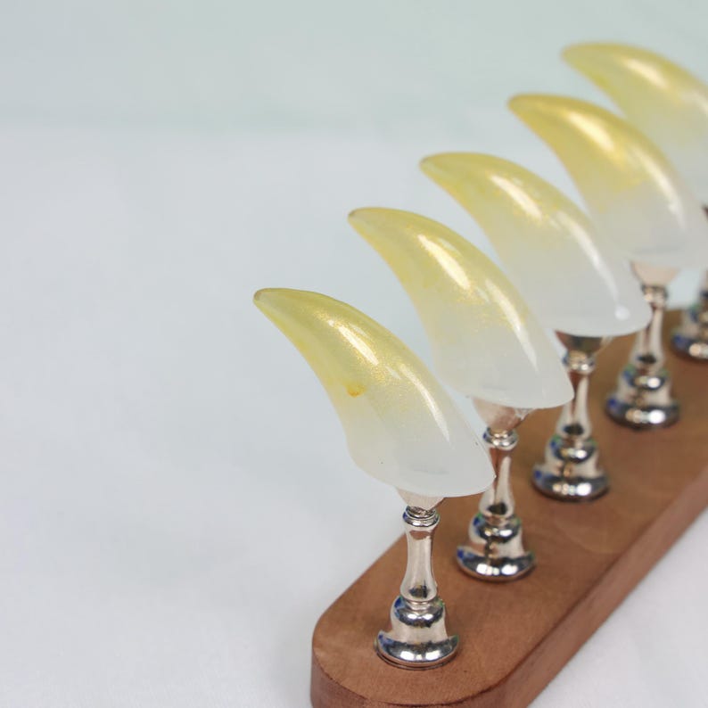 May include: A set of six decorative nail art display stands. Each stand features a clear, curved nail with a yellow gradient, mounted on a silver-colored pedestal. The stands are arranged on a wooden base, creating a unique display.