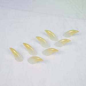 May include: A collection of eight translucent, claw-shaped objects. The claws transition from a pale yellow at the tips to a clear, colorless base. They are arranged on a white surface.