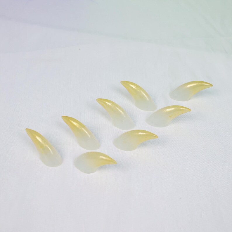 May include: A collection of eight translucent, claw-shaped objects. The claws transition from a pale yellow at the tips to a clear, colorless base. They are arranged on a white surface.