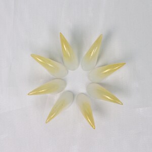 May include: Eight long, pointed, artificial fingernails arranged in a circular pattern. The nails transition from a white base to a yellow-gold tip, creating a gradient effect. The nails are set against a white background.