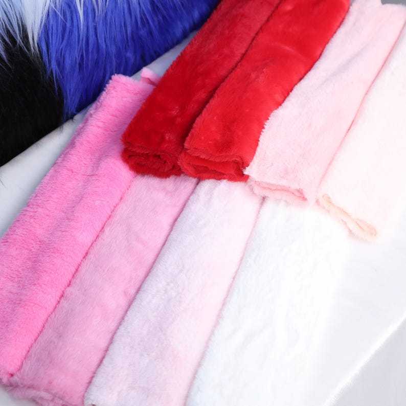 May include: A collection of colorful faux fur fabric in shades of red, pink, and white. The plush material is arranged in folded layers, showcasing the soft texture and vibrant colors. Blue and black fur are visible in the background.