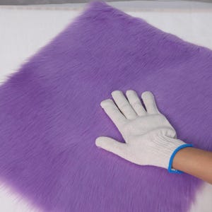 May include: A square of vibrant purple faux fur. The plush material has a soft, textured appearance. A white glove with a blue trim is touching the fur, highlighting its texture.