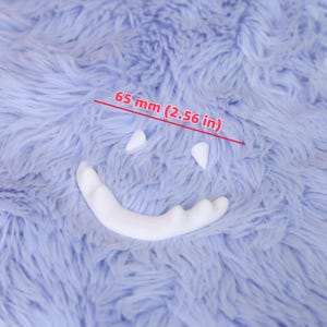 Puede incluir: Un diseño de cara sonriente blanca con dos ojos triangulares y una boca curva, sobre un fondo azul claro y esponjoso. El diseño mide 6,5 cm de ancho.