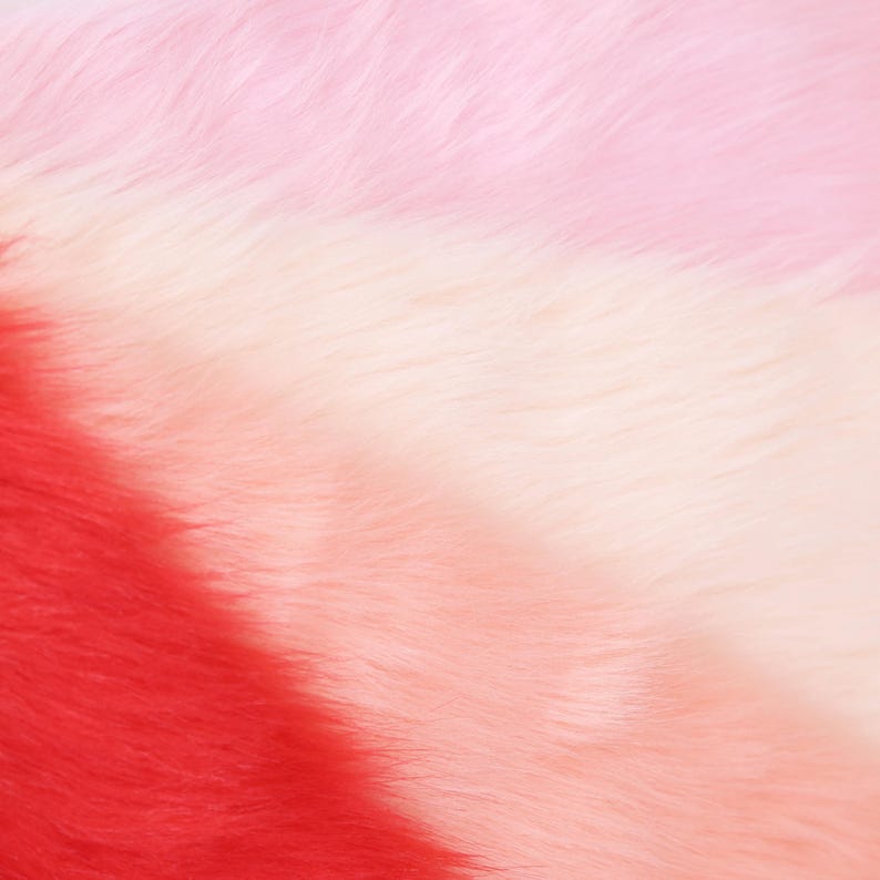 May include: Close-up of a faux fur fabric with a gradient of colors. The colors transition from red to light pink and white, creating a soft, textured appearance. The fur fibers are visible, adding depth and dimension.