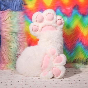 Op de afbeelding: Een pluche witte kattenpoot met roze kussentjes en klauwen. De poot is gemaakt van zacht, pluizig materiaal. De achtergrond heeft kleurrijk, regenboogkleurig imitatiebont.