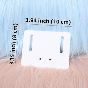 May include: A white rectangular product display card with two vertical slots and two small holes. The card measures 10 cm wide and 8 cm tall. The background is a mix of light blue and peach-coloured faux fur.