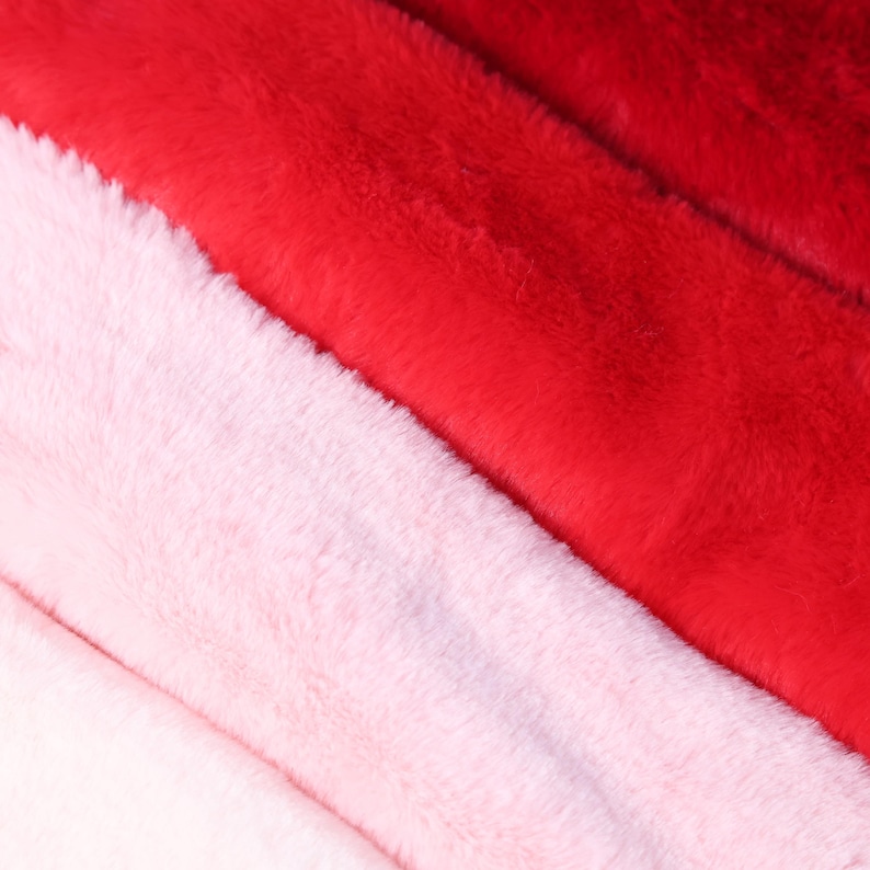May include: Close-up of three sections of faux fur fabric in shades of red and pink. The colors transition from a light pink on the left, to a medium pink in the center, and a vibrant red on the right. The fur has a soft, plush texture.