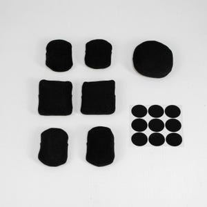 May include: A collection of six black fabric-covered pads in various shapes, including rectangular and rounded forms, alongside one larger circular black pad and a square sheet containing nine small black circular adhesive stickers.