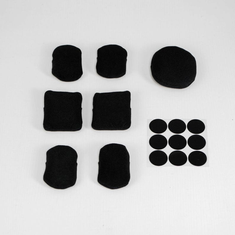 May include: A collection of six black fabric-covered pads in various shapes, including rectangular and rounded forms, alongside one larger circular black pad and a square sheet containing nine small black circular adhesive stickers.