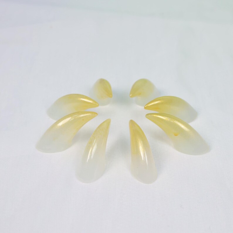 May include: A collection of eight artificial fingernails, arranged in a circular pattern. The nails are a gradient of white to yellow, with a glossy finish. The tips are a vibrant yellow, while the base is a translucent white.