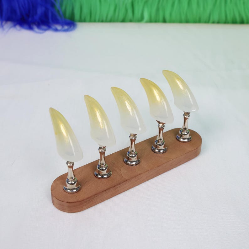 May include: Five yellow and white claw-shaped objects on silver stands, mounted on a wooden base. The claws have a gradient effect, transitioning from yellow to white. The wooden base is rectangular and has a natural wood finish.
