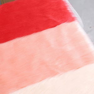 May include: Close-up of three sections of faux fur fabric in red, peach, and cream colors. The fur has a soft, textured appearance, with the fibers lying in the same direction. The fabric is likely used for crafts, costumes, or home decor projects.