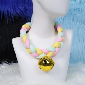 Op de afbeelding: Een pastelkleurige gevlochten halsband met een grote gouden bel. De halsband is gemaakt van zachte, geweven stof in tinten roze, geel en mintgroen. De bel is rond en glanzend en reflecteert het licht.