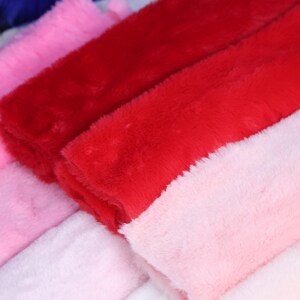 May include: Close-up of several plush Santa hat cuffs in various shades of red and pink. The soft, fuzzy texture of the fabric is visible, with a focus on the vibrant colors and the material's tactile quality. The hats are likely for the holiday season.