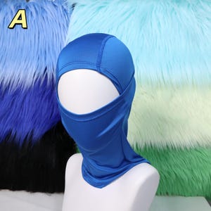 Op de afbeelding: Een felblauwe bivakmuts, ontworpen om het hoofd en de nek te bedekken, met een opening voor de ogen en mond. De bivakmuts wordt getoond op een witte mannequinhoofd. De achtergrond heeft kleurrijk imitatiebont in blauw- en groentinten.