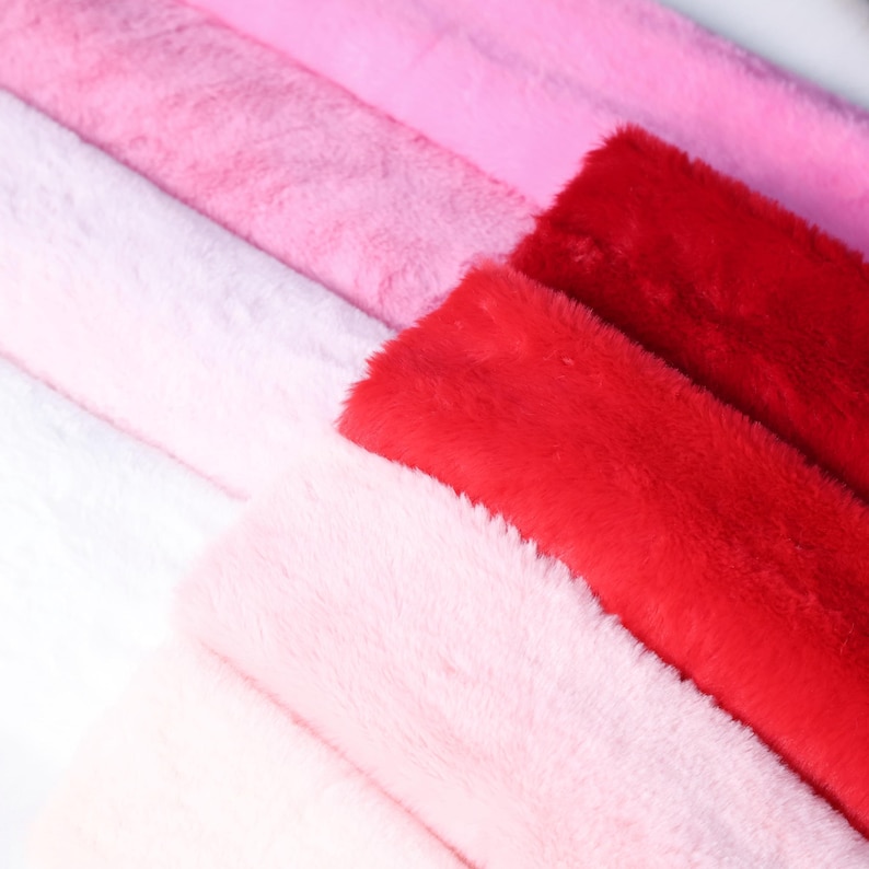 May include: A close-up of several rolls of faux fur fabric in various shades of pink and red. The colors range from light pink to a vibrant red, with a soft, fluffy texture. The fabric is arranged in diagonal rows.